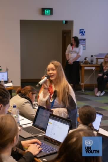 Emma speaking at an UNYouth event with a Māori flag wrapped around her