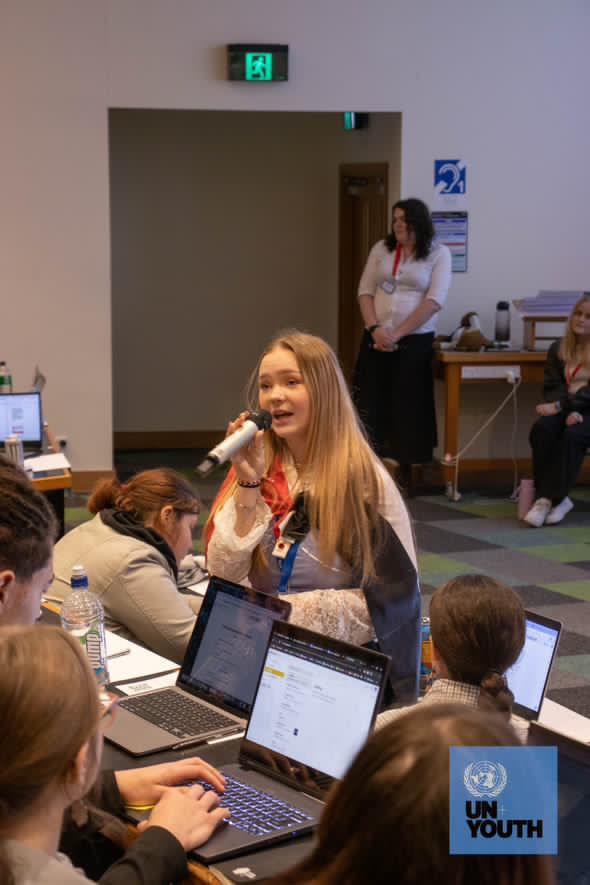 Emma speaking at an UNYouth event with a Māori flag wrapped around her